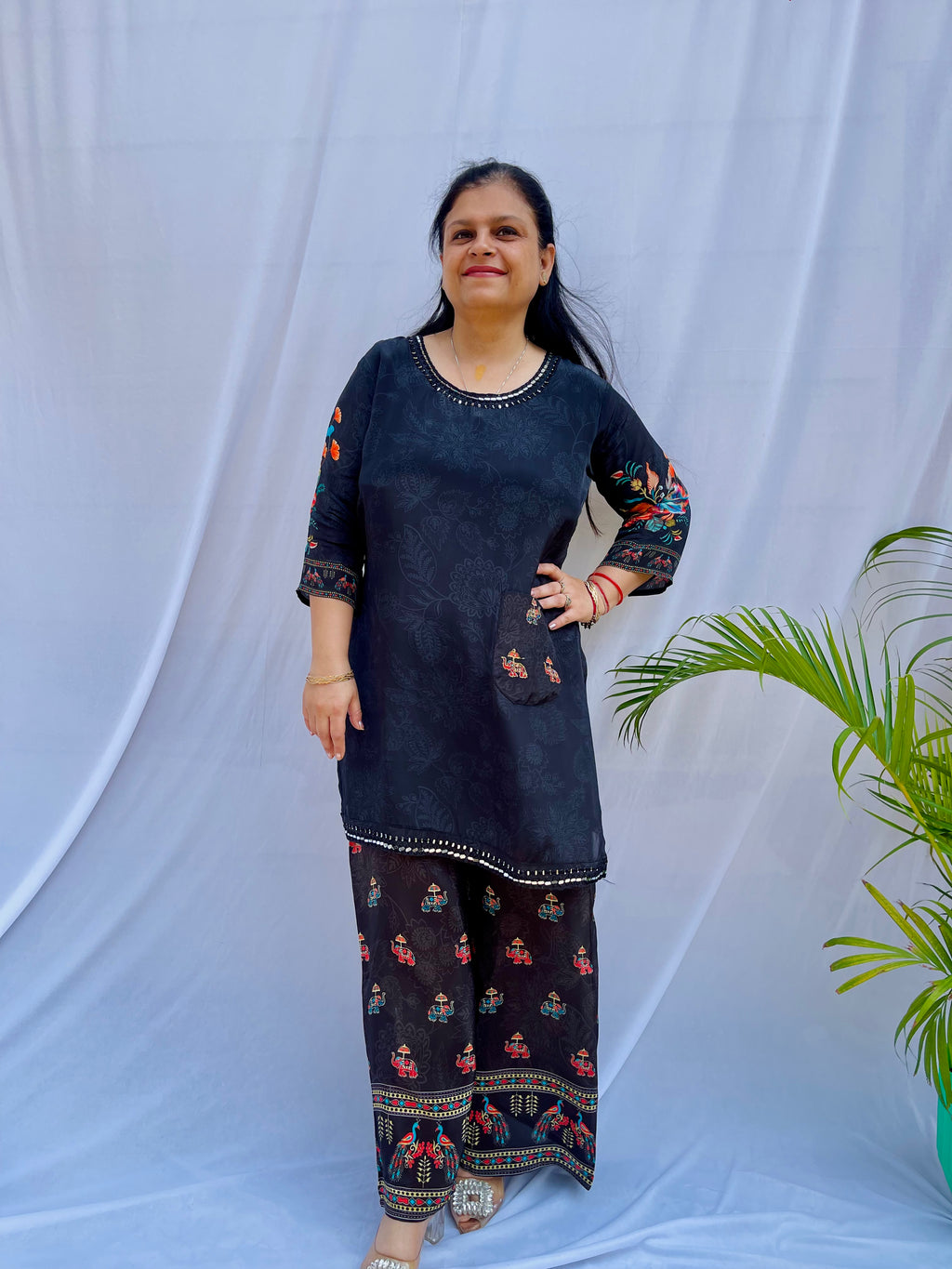 Model posing in a midnight black tunic with floral embroidered sleeves and wide-leg trousers featuring traditional elephant motifs.