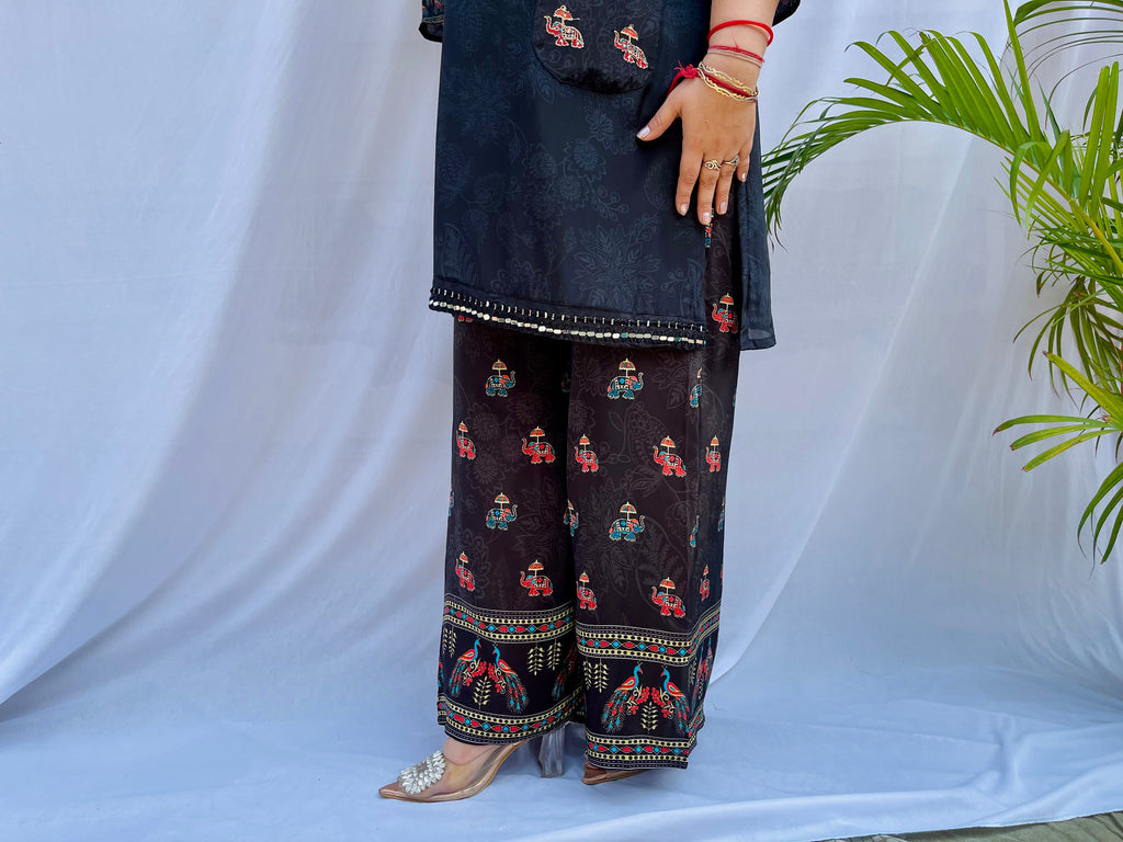 Model posing in a midnight black tunic with floral embroidered sleeves and wide-leg trousers featuring traditional elephant motifs.